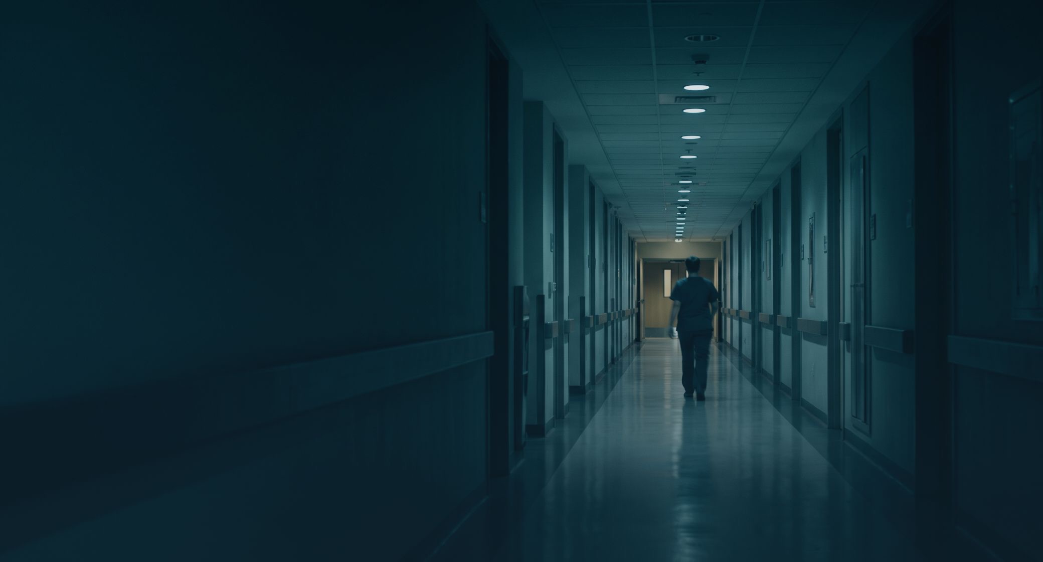 An empty hospital corridor at night, a single figure walking away in the far distance.